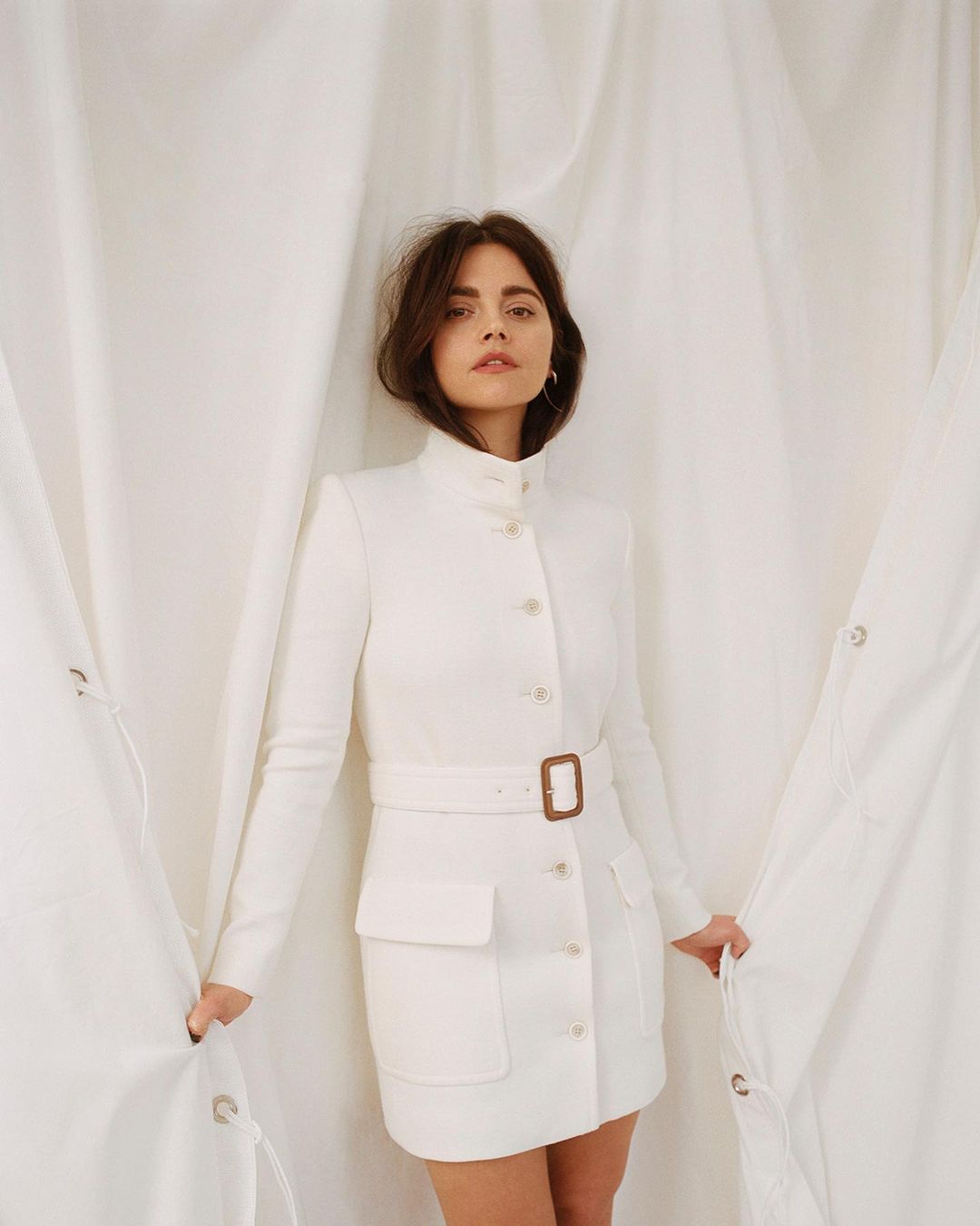 A brunette woman in a white belted dress with a high collar poses in front of a white fabric backdrop.