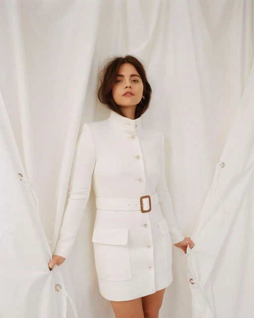 A brunette woman in a white belted dress with a high collar poses in front of a white fabric backdrop.