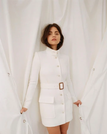 A brunette woman in a white belted dress with a high collar poses in front of a white fabric backdrop.