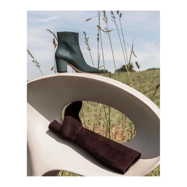 Two leather boots, one brown, one green, on a modern white chair outdoors with field and blue sky in the background. Fashion still life.