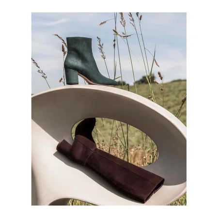 Two leather boots, one brown, one green, on a modern white chair outdoors with field and blue sky in the background. Fashion still life.