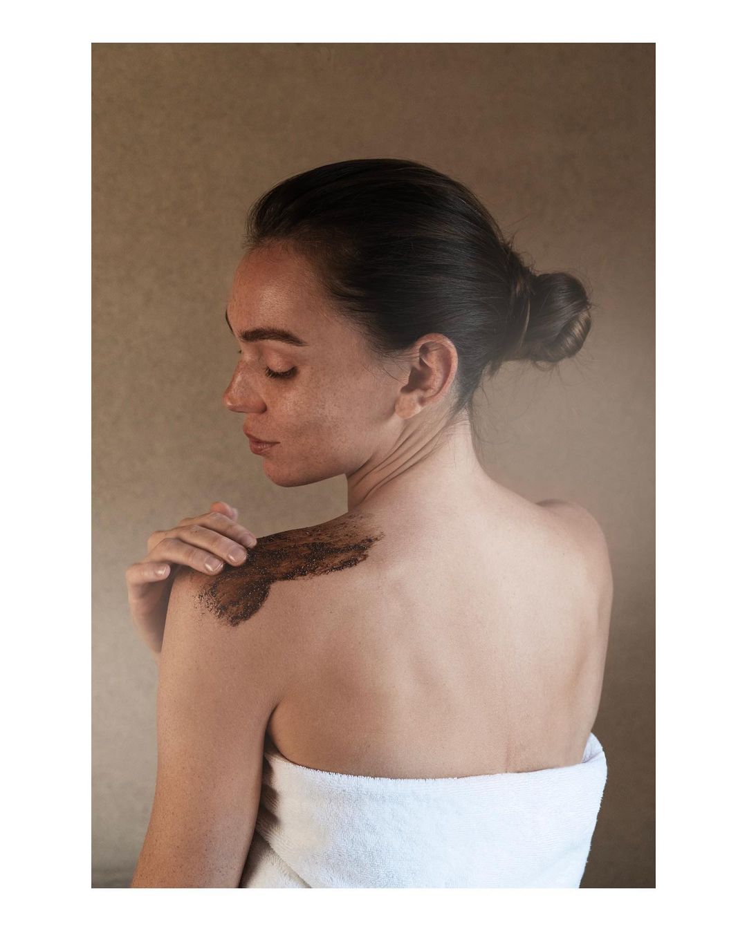 Woman with dark hair and a towel is seen from the rear, applying a body scrub to her shoulder in a close-up shot against a neutral background.