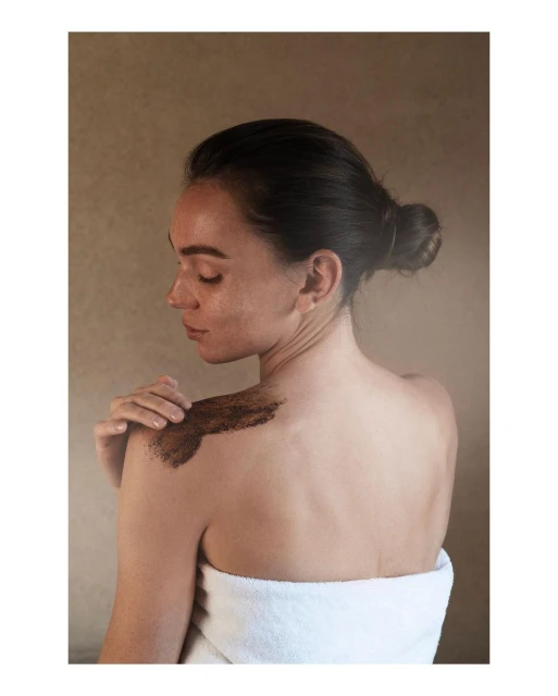 Woman with dark hair and a towel is seen from the rear, applying a body scrub to her shoulder in a close-up shot against a neutral background.