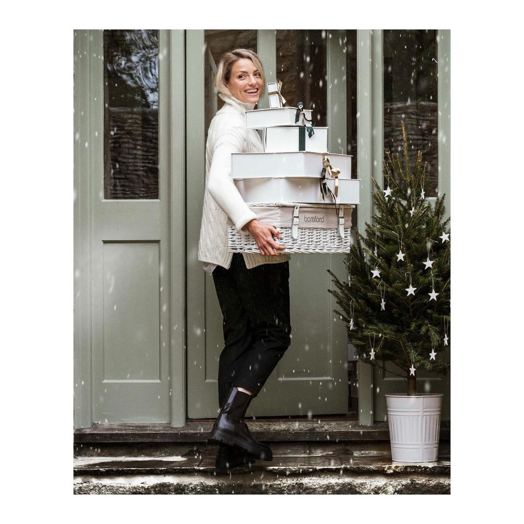 Smiling woman carries stack of white gift boxes near Christmas tree at house entrance with grey-green door during snow fall.