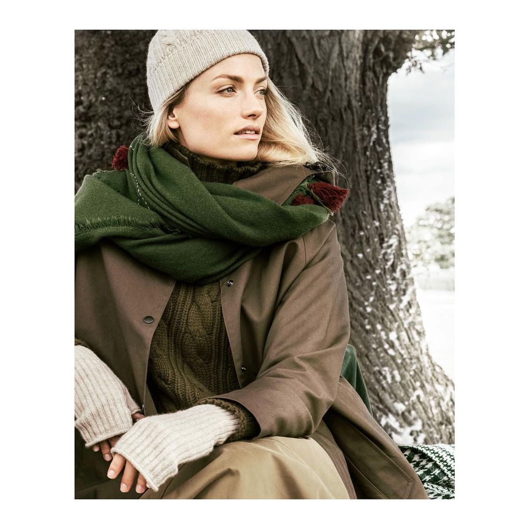 Woman in a beanie and scarf poses outdoors next to a tree in an outdoor setting on a winter day, showcasing fashion apparel.