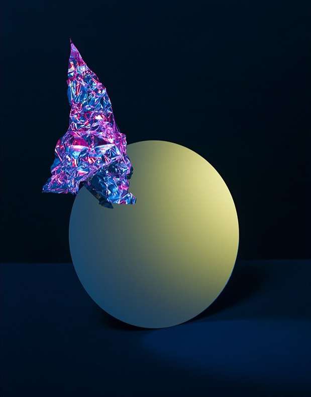Abstract still life featuring a metallic, crumpled form balanced on a matte sphere against a dark blue backdrop in studio lighting.