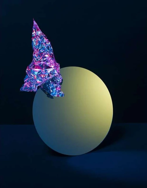 Abstract still life featuring a metallic, crumpled form balanced on a matte sphere against a dark blue backdrop in studio lighting.