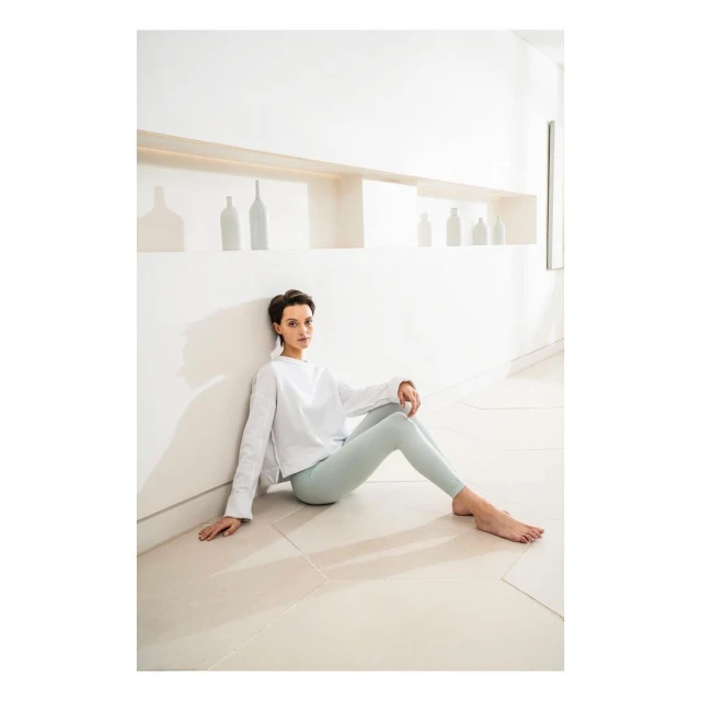 Woman in white top and light green leggings sits barefoot indoors, leaning against a white wall with decorative bottles on a shelf.
