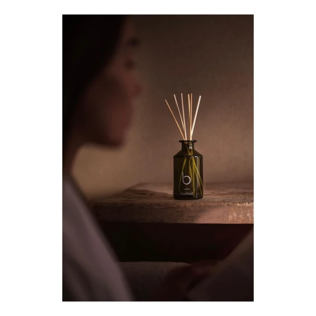 Reed diffuser in a dark green glass bottle on a weathered wooden surface with a blurred female figure in the foreground.