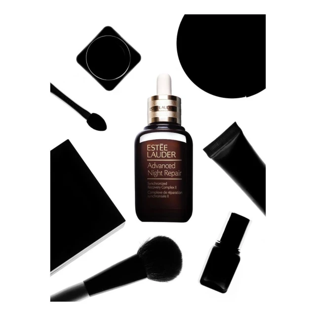 Estee Lauder Advanced Night Repair serum with dropper surrounded by beauty products in a still-life composition on white.