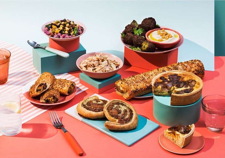 A variety of foods, including pastries, falafel, salad, and shredded chicken, are arranged on plates and platforms against a colorful background.