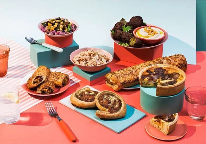 A variety of foods, including pastries, falafel, salad, and shredded chicken, are arranged on plates and platforms against a colorful background.