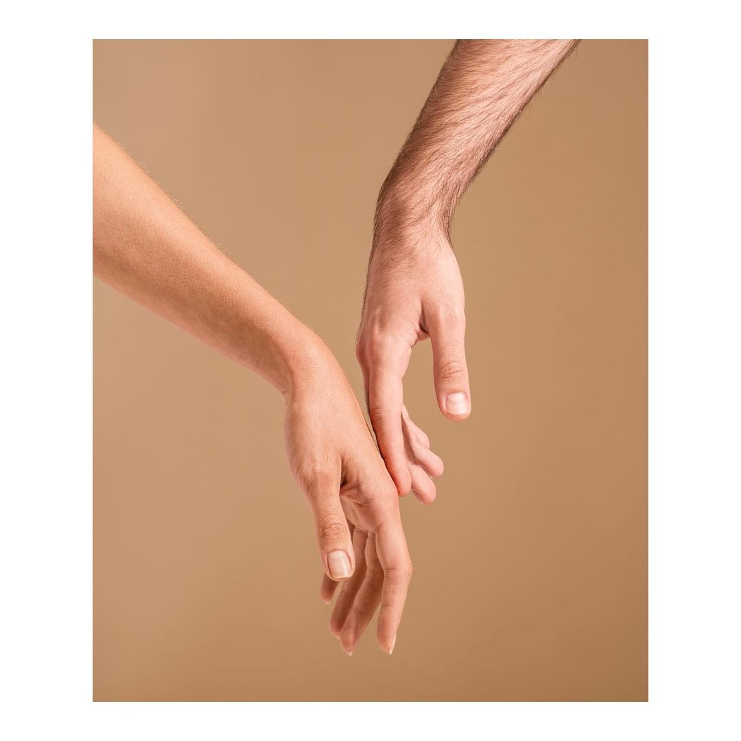 Close-up of a woman's arm and a man's arm reaching towards each other on a neutral background, conveying a sense of connection and intimacy.