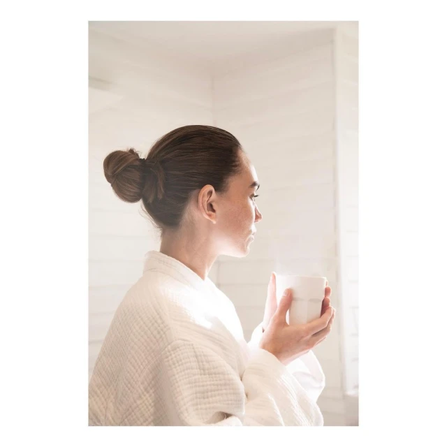 Woman in a white robe looks out of a window while holding a mug in a bright room with white walls and a light-filled background.