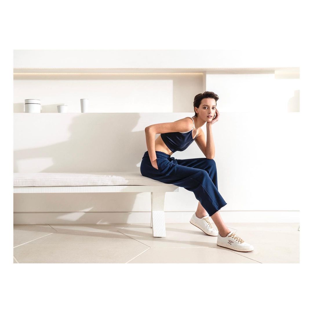 A slender woman with cropped hair poses on a white bench wearing a navy crop top and pants, with white sneakers in a brightly lit setting.
