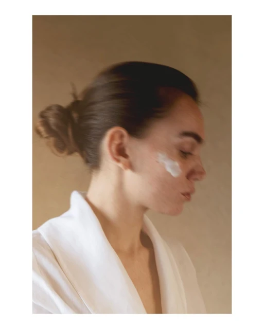 Close-up profile of a young woman wearing a white robe and facial cream on her cheek, with a blurred background and soft studio lighting.