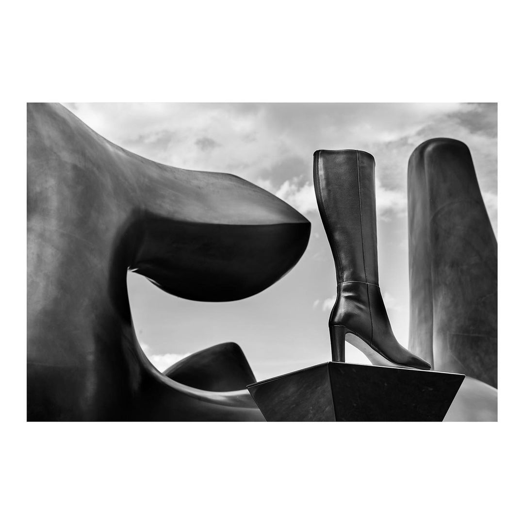 Black and white image of a high-heeled leather boot displayed on a geometric structure against a partly cloudy sky, creating a surreal effect.