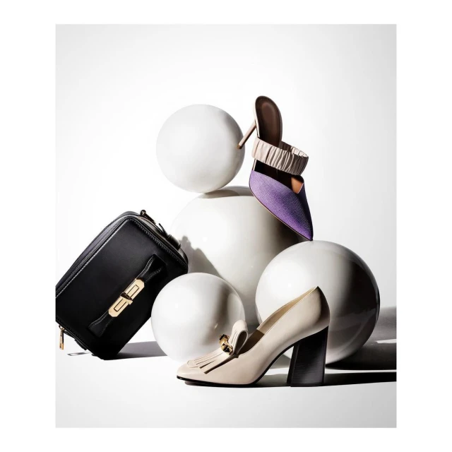 Still life of a black purse, beige loafer, and a purple heeled shoe arranged with spherical white objects on a white background.