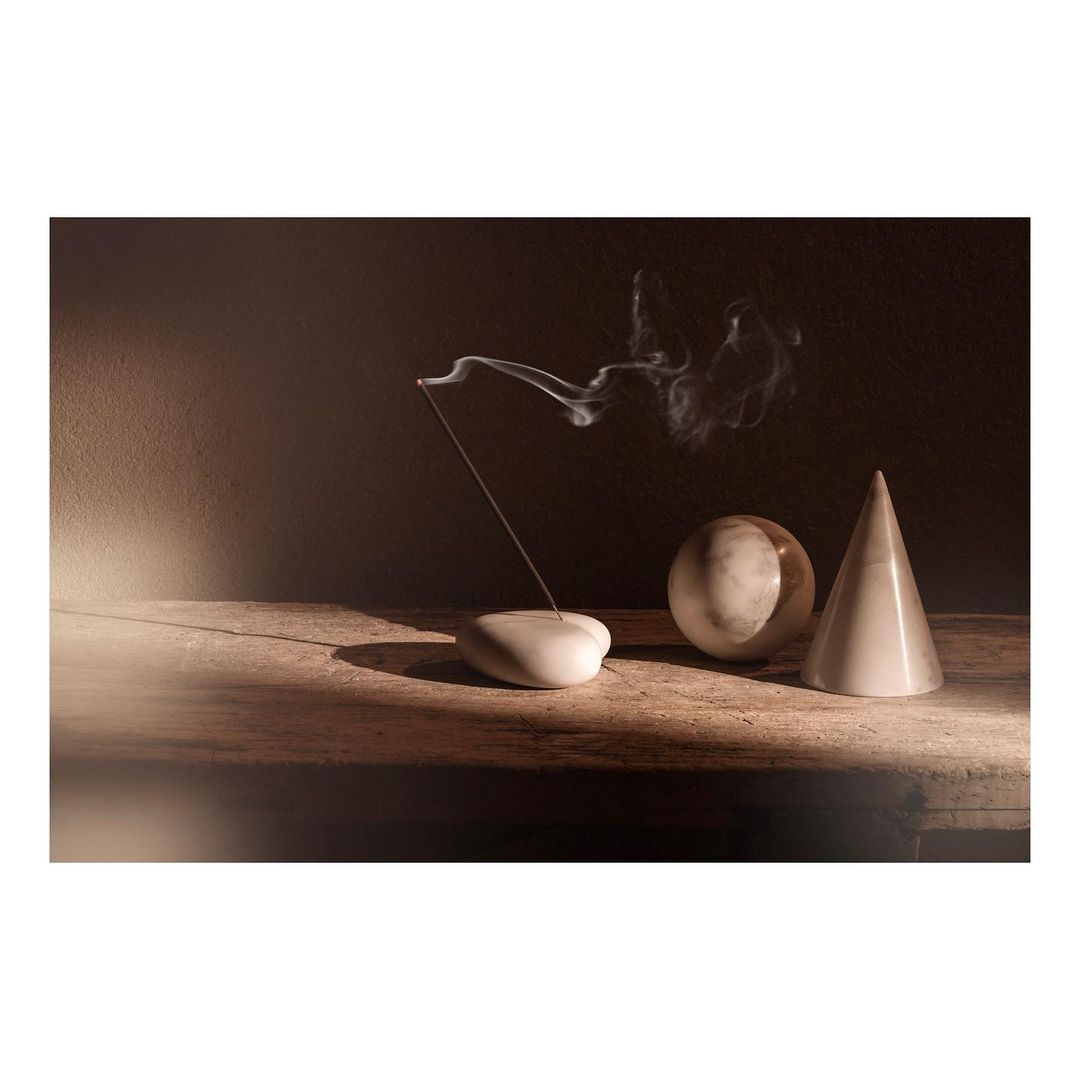 Still life with smoking incense stick, smooth white pebble holder, marble sphere and cone on a rustic wood surface against a dark background.