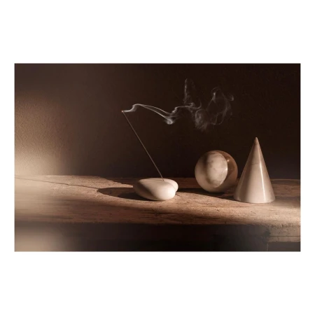 Still life with smoking incense stick, smooth white pebble holder, marble sphere and cone on a rustic wood surface against a dark background.