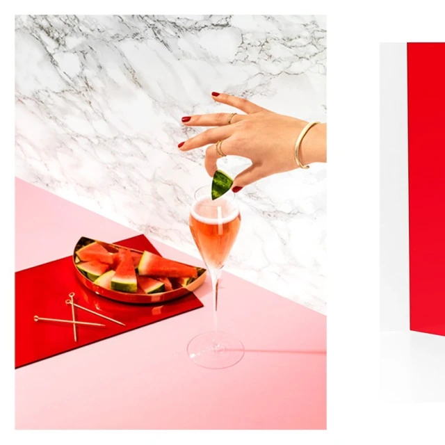 Still life shot of a person adding a watermelon slice to a champagne flute, with a tray of watermelon and gold cocktail sticks on a pink surface.