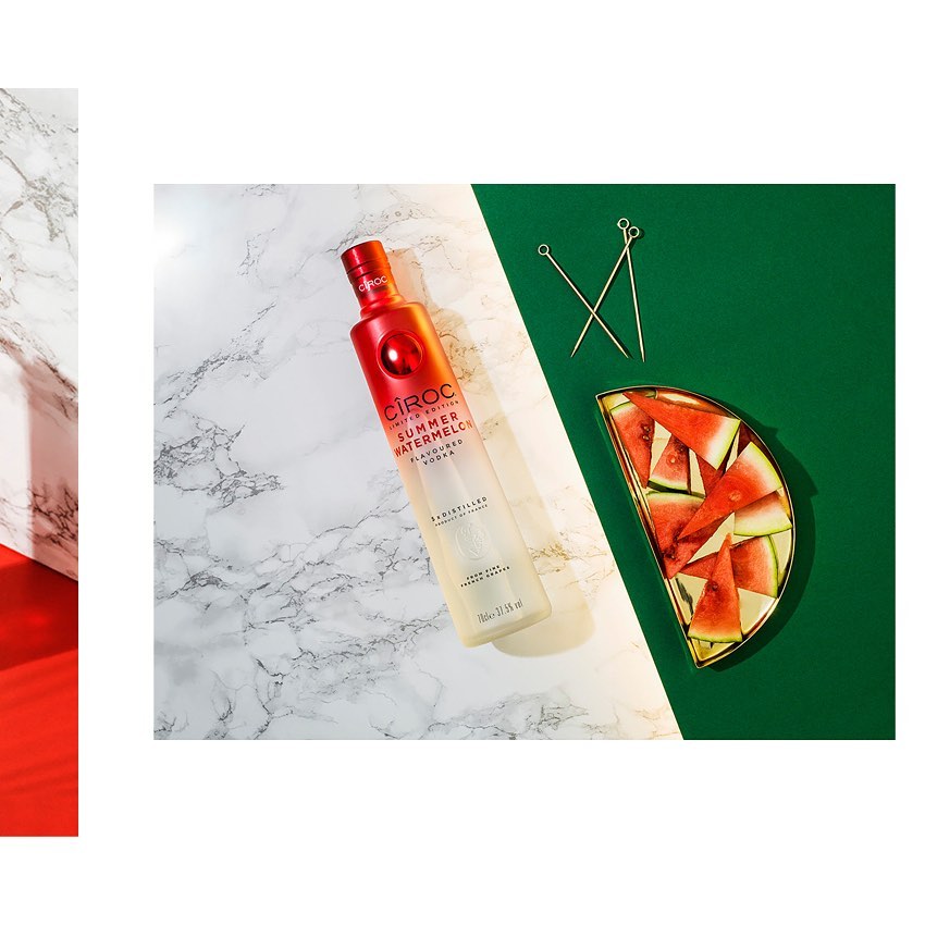 Ciroc Summer Watermelon vodka bottle angled on a marble surface with watermelon slices in a semi-circular gold dish on green backdrop.