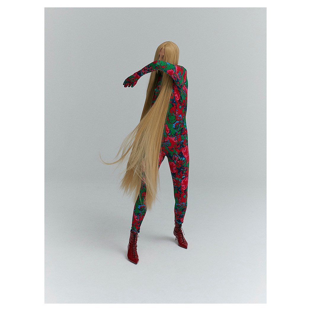 Fashion-forward figure in floral jumpsuit with cascading blonde hair strikes a dynamic pose in a stark studio setting, highlighted by red heels.