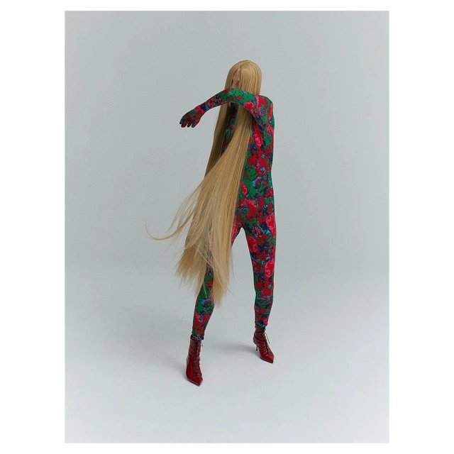Fashion-forward figure in floral jumpsuit with cascading blonde hair strikes a dynamic pose in a stark studio setting, highlighted by red heels.