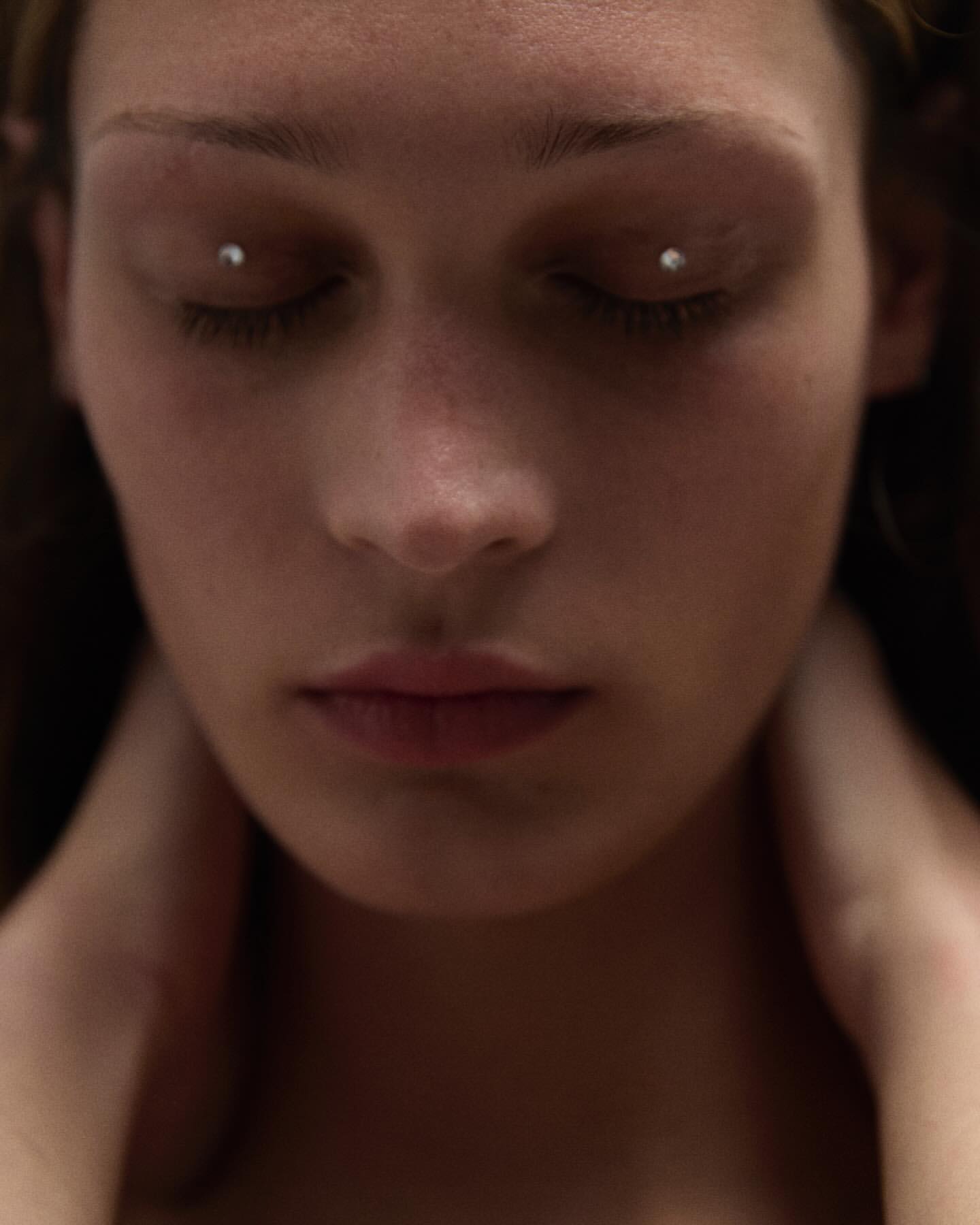 Close-up portrait of a woman with closed eyes and eye piercings, focusing on her facial features and neutral expression, indoors.
