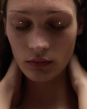 Close-up portrait of a woman with closed eyes and eye piercings, focusing on her facial features and neutral expression, indoors.