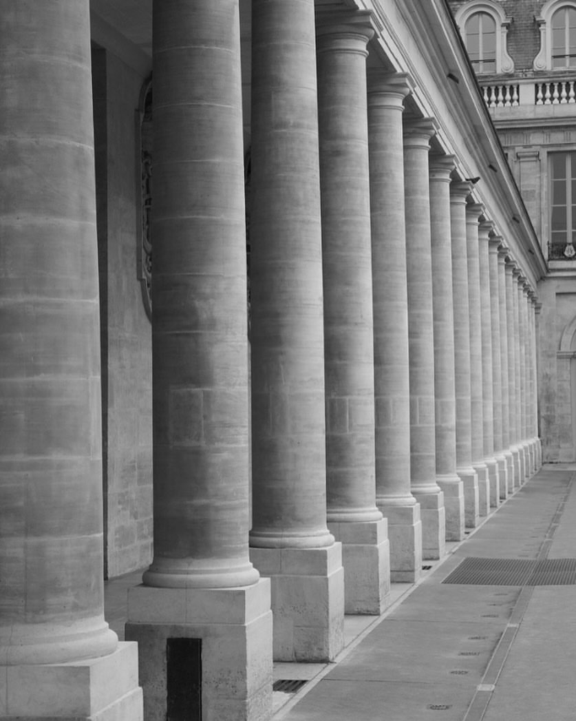Black and white architectural photograph features a row of tall, evenly spaced stone pillars in a building colonnade under natural light.