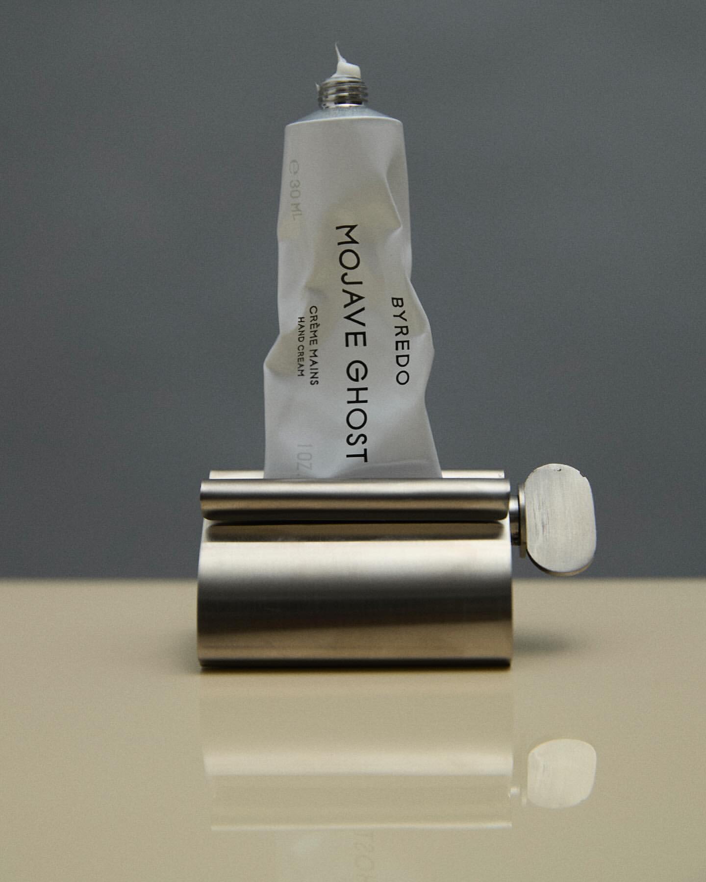 Byredo Mojave Ghost hand cream in a metal dispenser on a beige surface with a grey background. Beauty and personal care product shot.