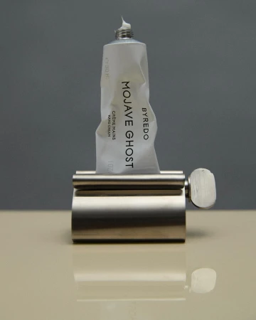 Byredo Mojave Ghost hand cream in a metal dispenser on a beige surface with a grey background. Beauty and personal care product shot.