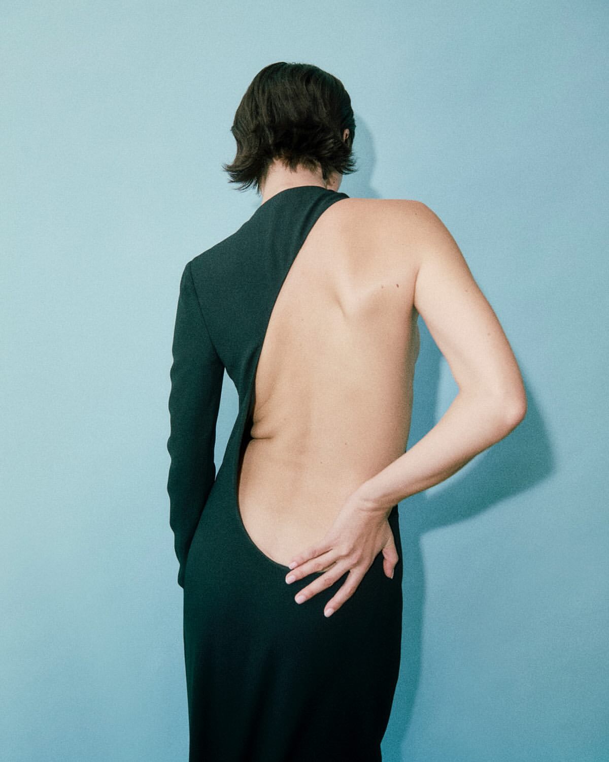 Adult woman in dark dress with exposed back poses against blue studio backdrop in a fashion portrait with natural, soft lighting.