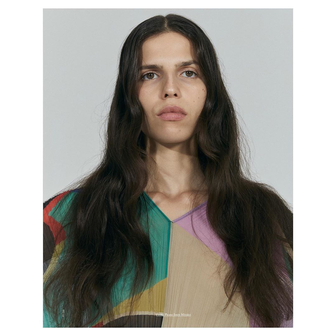 Close-up studio portrait of a person with long brown hair wearing a colorful top with a v-neck against a neutral background.