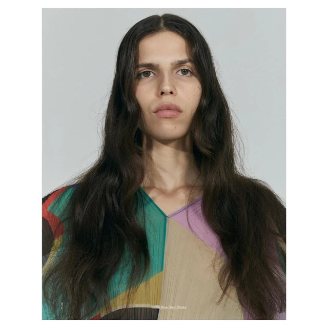 Close-up studio portrait of a person with long brown hair wearing a colorful top with a v-neck against a neutral background.