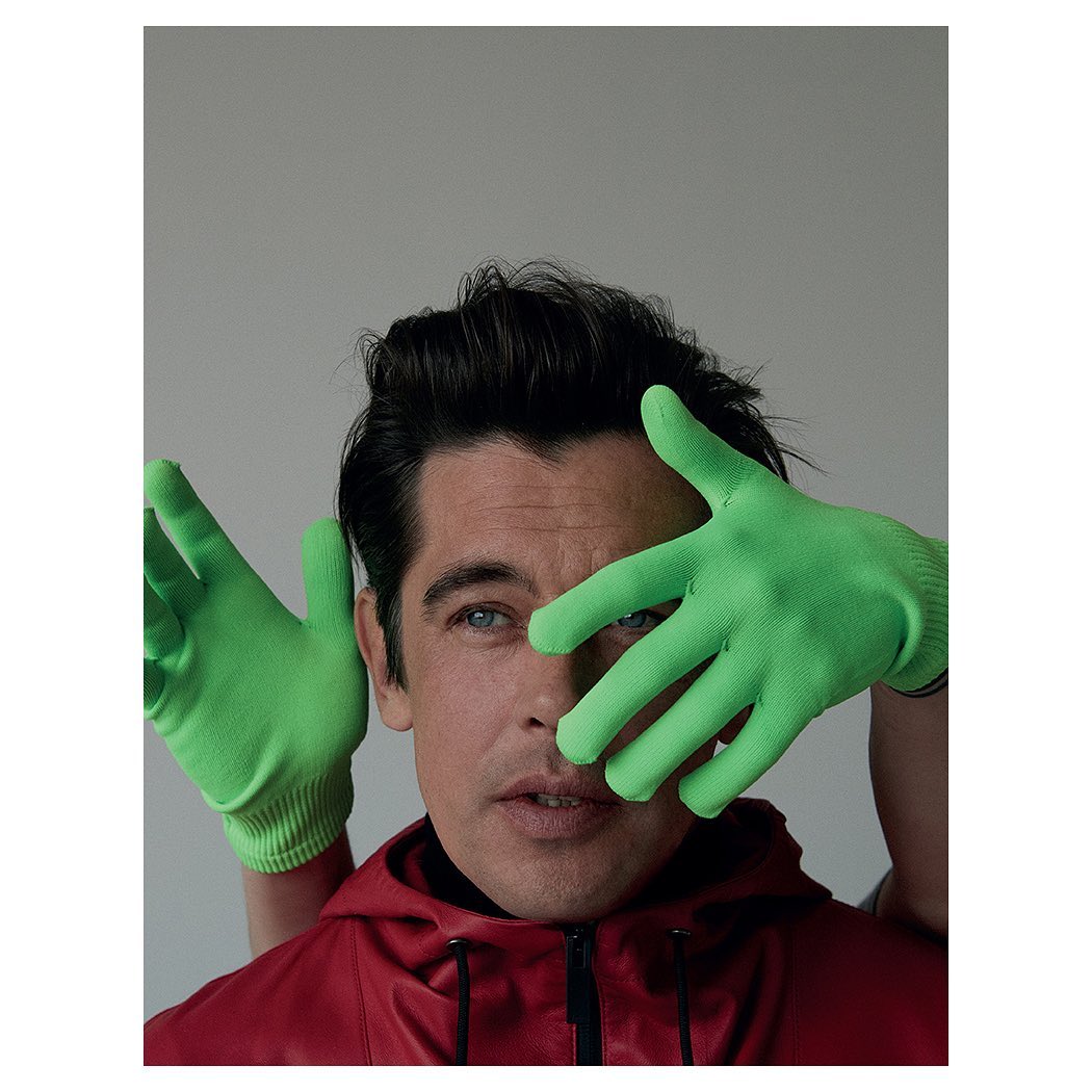 Close-up portrait of a man with dark hair and blue eyes, wearing a red jacket and bright green gloves, against a plain grey background.