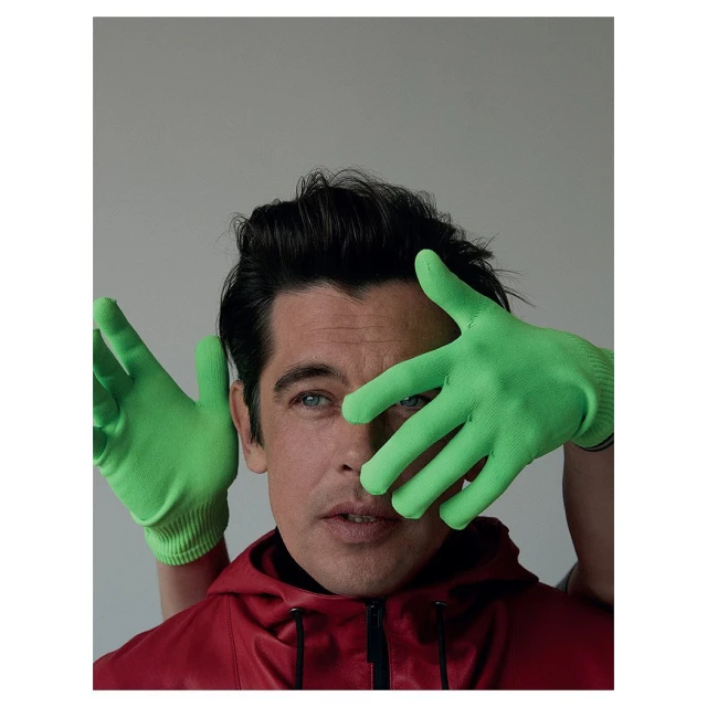 Close-up portrait of a man with dark hair and blue eyes, wearing a red jacket and bright green gloves, against a plain grey background.