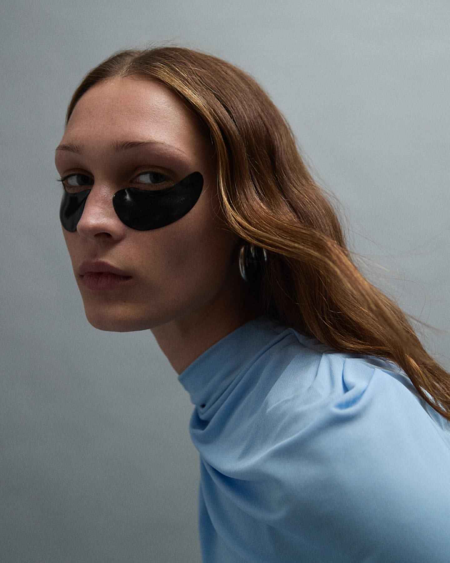 Close-up of an adult female with brown hair wearing black eye mask patches and a light blue draped top against a light gray background.