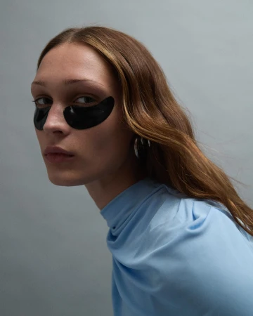Close-up of an adult female with brown hair wearing black eye mask patches and a light blue draped top against a light gray background.