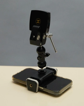Close-up studio shot of black smartphone attached to an adjustable metal tripod stand with a Chanel logo on the phone's back.