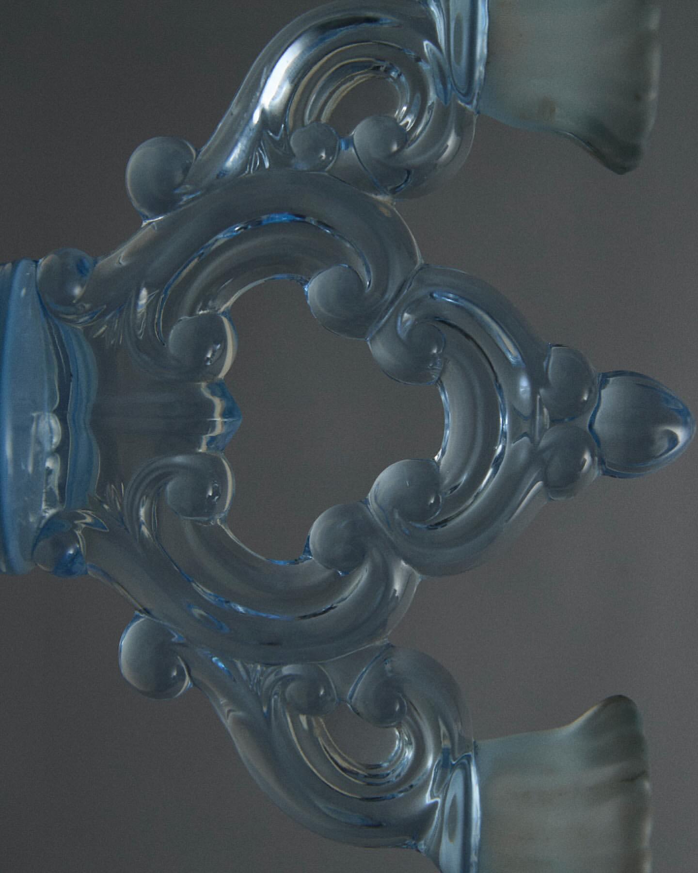Close-up of an ornate pale-blue glass object with curved flourishes and embellishments, all set against a muted gray background.