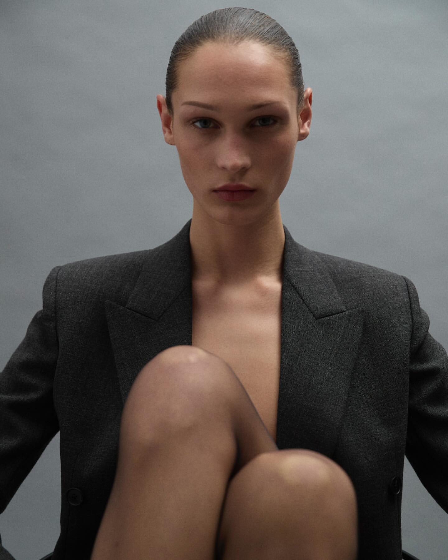 Portrait of a woman with slicked-back hair wearing a grey blazer and black tights against a plain grey studio background.