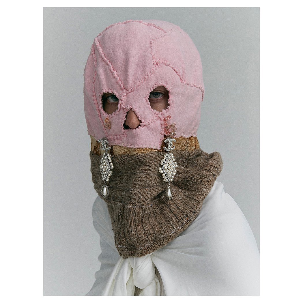 Fashion portrait featuring a person wearing a pink patchwork balaclava with cut-out eye and mouth holes, paired with pearl earrings.