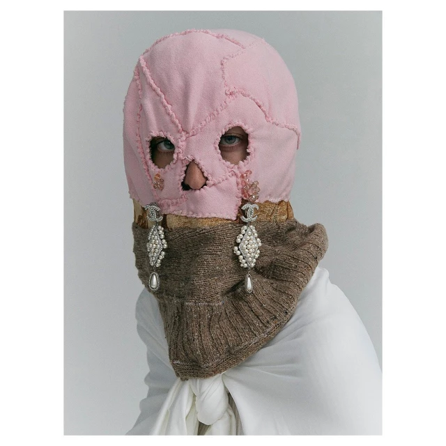 Fashion portrait featuring a person wearing a pink patchwork balaclava with cut-out eye and mouth holes, paired with pearl earrings.