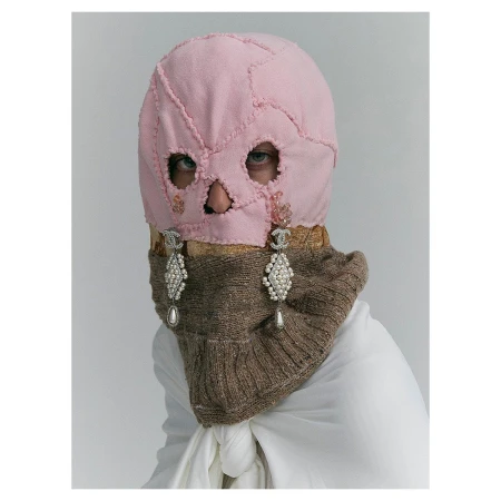 Fashion portrait featuring a person wearing a pink patchwork balaclava with cut-out eye and mouth holes, paired with pearl earrings.