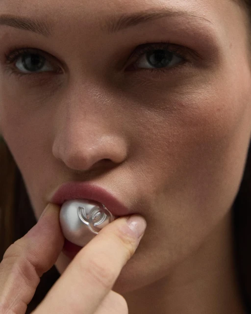 Close-up of a fair-skinned woman holding a pearl-like orb with the Chanel logo between her lips and finger, beauty product.