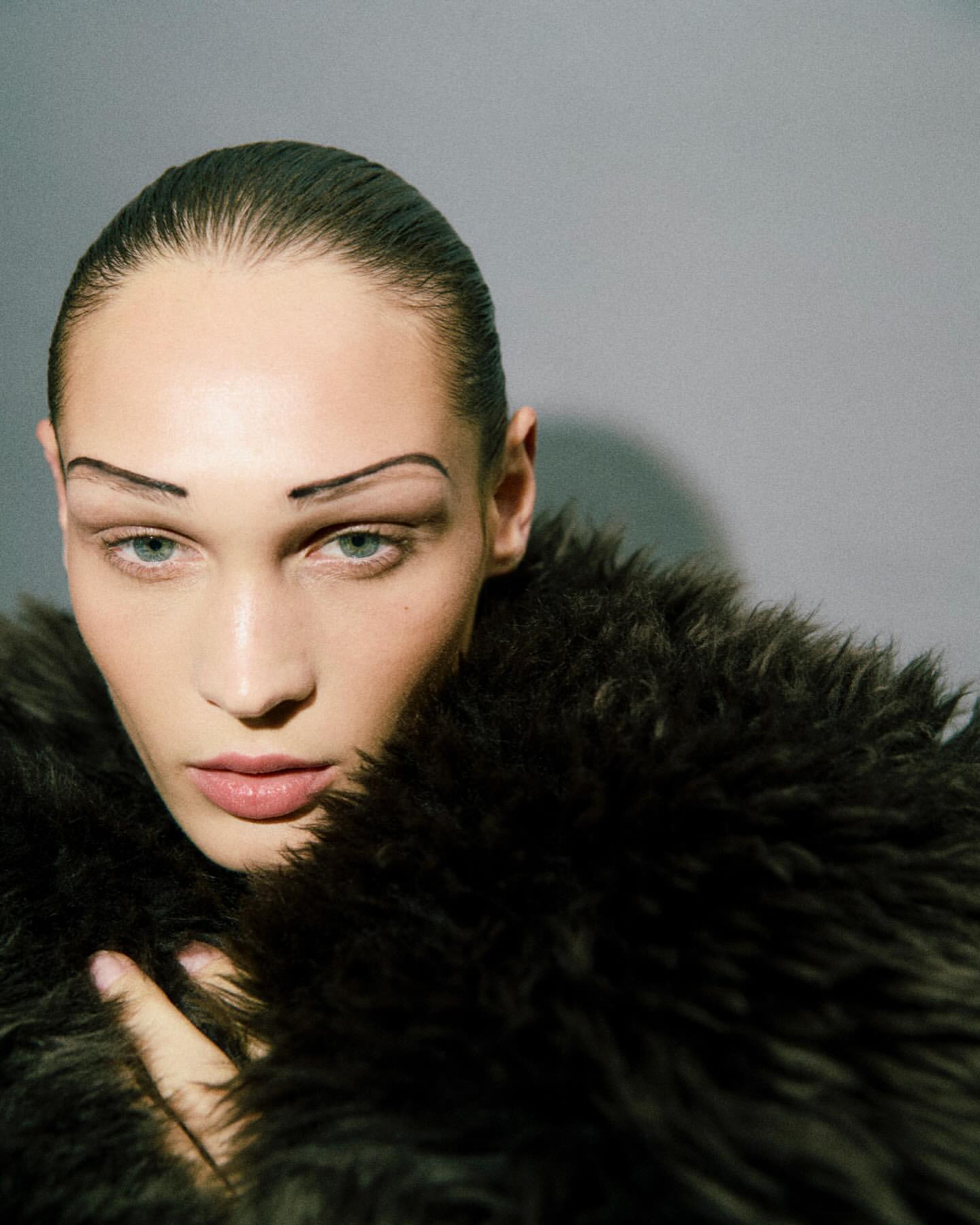 Close-up shot of a woman with dark hair and heavy makeup, wearing a fur coat against a neutral grey background in a beauty editorial.