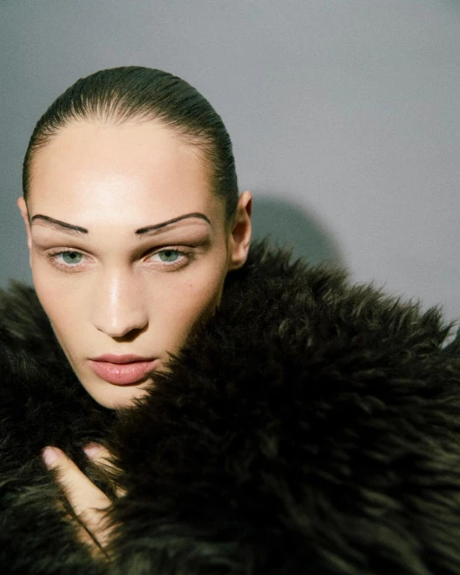 Close-up shot of a woman with dark hair and heavy makeup, wearing a fur coat against a neutral grey background in a beauty editorial.