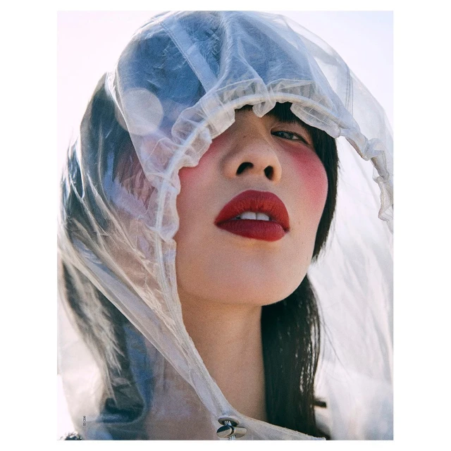 Close-up portrait of a young woman with bold red lipstick and blush, wearing a transparent plastic hooded raincoat.